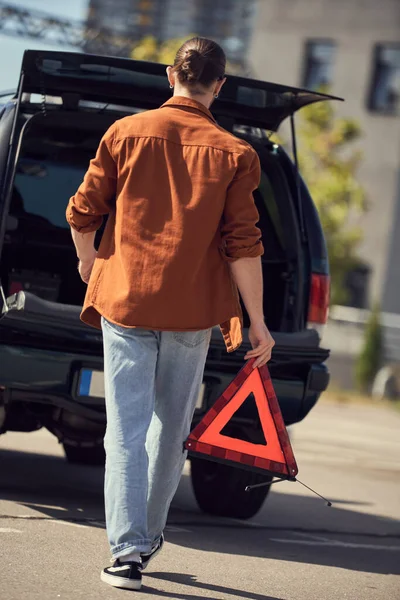 Vista trasera del joven hombre con estilo en traje urbano caminando a su coche con triángulo de advertencia en las manos - foto de stock