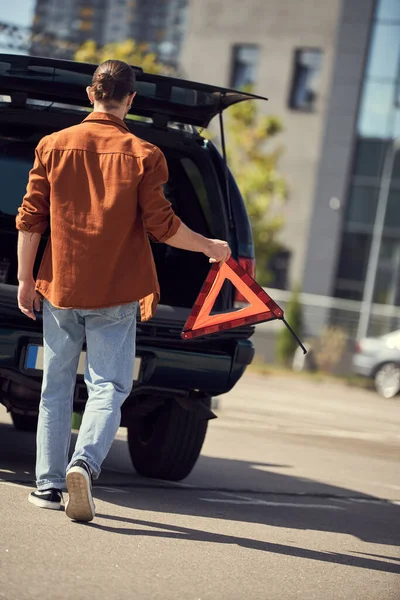 Visão traseira do jovem em camisa marrom andando até seu carro com triângulo de advertência nas mãos, motorista sexy — Fotografia de Stock