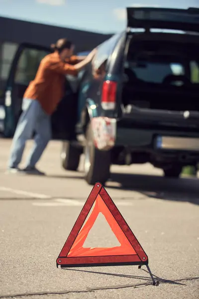 Photo floue du jeune homme en tenue urbaine stylée debout à côté de sa voiture, triangle d'avertissement — Photo de stock