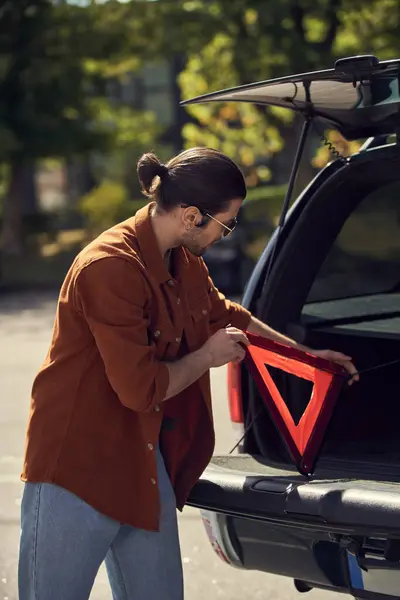 Tiro vertical del hombre con estilo con gafas de sol poniendo triángulo de advertencia en el maletero de su coche, conductor sexy - foto de stock