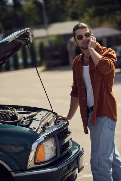 Colpo verticale del conducente maschio che parla per telefono con il suo assicuratore vicino al cofano motore aperto di auto rotta — Foto stock