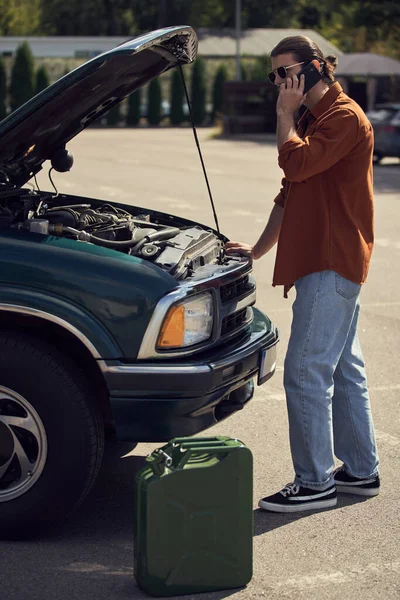 Vertical shot of stylish man standing next to car and petrol canister talking by phone with insurer — Stock Photo