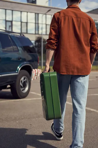 Обрезанный вид молодого стильного человека в джинсах и рубашке, держащего канистру с бензином, идущего к своей машине — стоковое фото