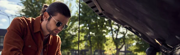 Modelo masculino sexy pensativo com óculos de sol e barba olhando atentamente para o seu motor de carro, banner — Fotografia de Stock