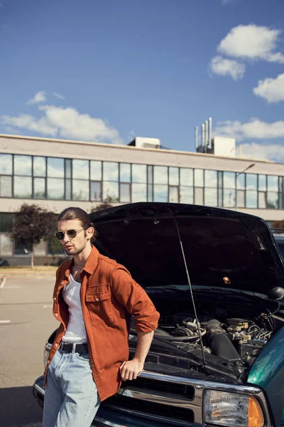 Vertical shot of attractive elegant man with dapper look standing near car with opened engine hood — Stock Photo