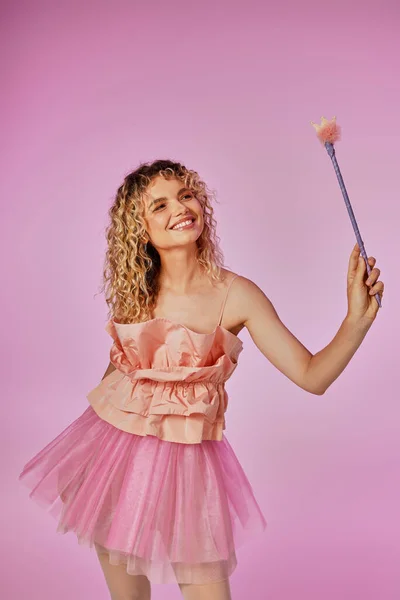 Cheerful tooth fairy in pink clothing with magic wand in hands looking away on pink backdrop — Stock Photo