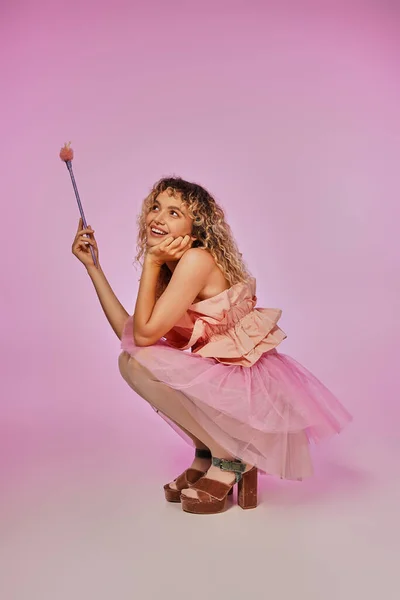 Jolie femme en costume de fée dentaire avec baguette magique dans les mains accroupi et posant sur fond rose — Photo de stock