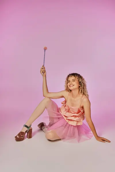 Gaie femme aux cheveux bouclés en costume de fée dent rose assis sur le sol et posant avec baguette magique — Photo de stock