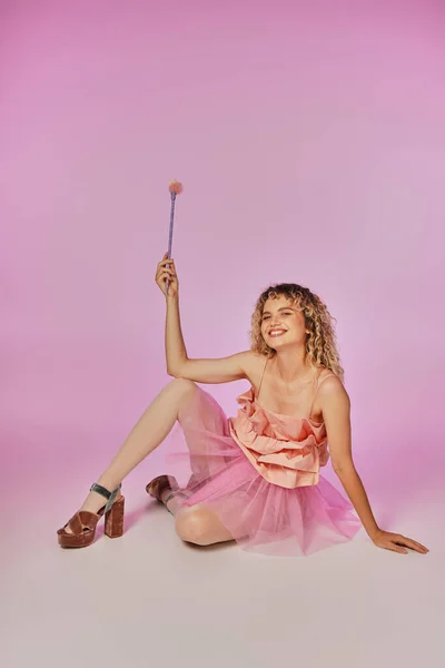 Cheerful curly haired woman in pink tooth fairy costume sitting on floor and posing with magic wand — Stock Photo