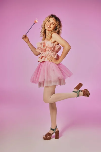 Веселая блондинка с вьющимися волосами в костюме зубной феи, стоящая на одной ноге на розовом фоне — стоковое фото