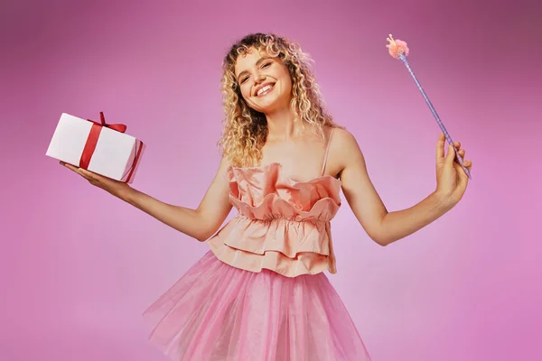 Усміхнена прекрасна жінка з кучерявим волоссям в рожевому костюмі тримає подарунок і чарівну паличку на рожевому тлі — стокове фото