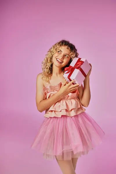 Mulher de cabelos encaracolados alegre em traje rosa olhando alegremente para a câmera e segurando presente perto do rosto — Fotografia de Stock
