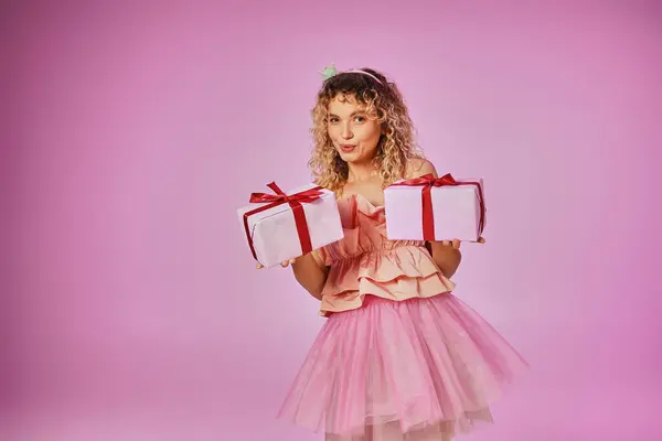 Femme blonde rêveuse en tenue rose de fée des dents montrant deux cadeaux sur la caméra sur fond rose — Photo de stock