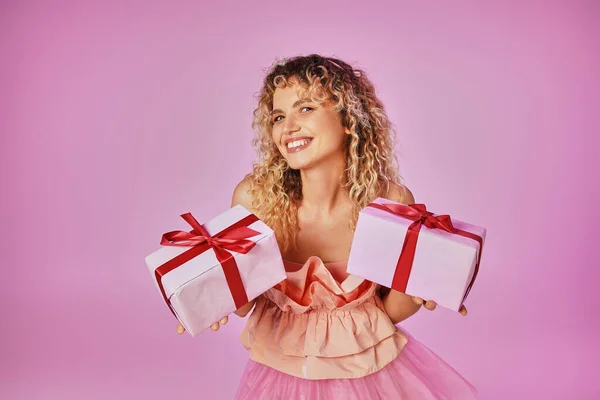 Alegre mujer de pelo rizado mirando felizmente a la cámara y sosteniendo dos regalos posando sobre fondo rosa - foto de stock