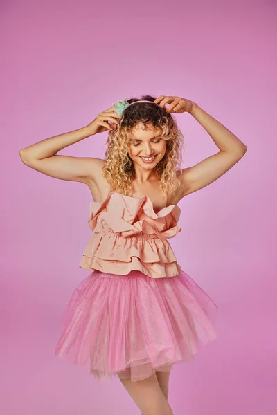 Encantadora hada de pelo rizado en traje rosa tocando su diadema y sonriendo en el fondo rosa - foto de stock