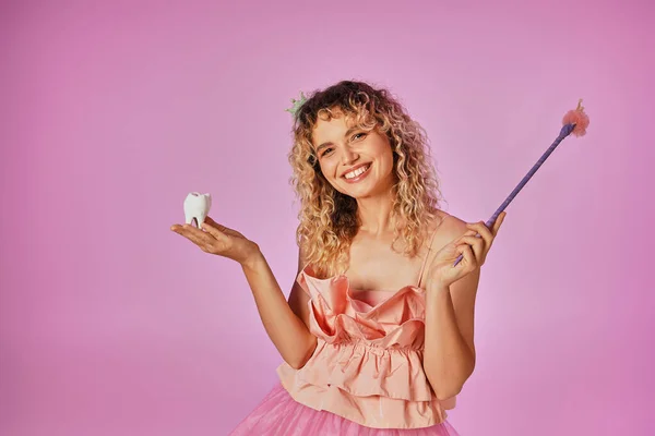 Joyous curly haired tooth fairy in pink costume and headband holding baby tooth and magic wand — Stock Photo