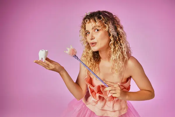 Lovely blonde woman in tooth fairy costume playfully casting spell on baby tooth with her magic wand — Stock Photo