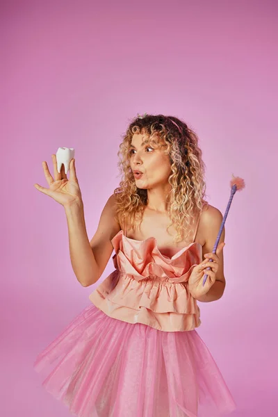 Amazed curly haired woman in tooth fairy attire with headband and magic wand looking at baby tooth — Stock Photo
