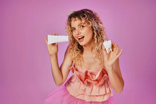 Усміхнена приваблива жінка в рожевому костюмі, що тримає зубну пасту та дитячий зуб на рожевому тлі — стокове фото