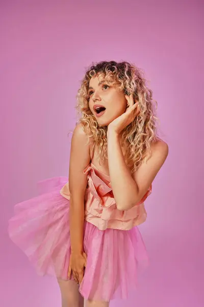 Encantadora mujer sorprendida en traje de hada diente rosa posando con la mano levantada a la mejilla y mirando hacia otro lado - foto de stock