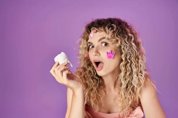 Portrait of amazed blonde woman with face stickers holding baby tooth and looking shocked at camera — Stock Photo
