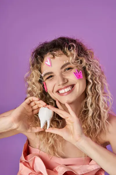 Веселая прекрасная женщина с вьющимися волосами и наклейками на лице, держащая молочный зуб и улыбающаяся в камеру — стоковое фото