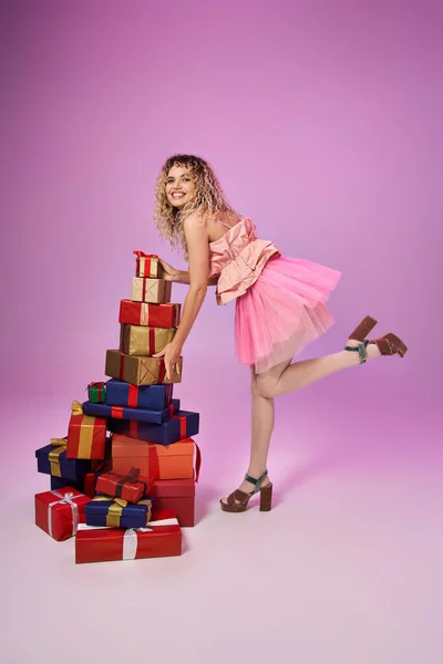 Mujer rubia feliz en traje de hada de dientes de pie en una pierna cerca de montón de regalos sobre fondo rosa - foto de stock