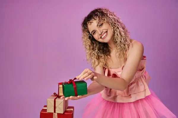 Alegre rubia en traje rosa sosteniendo uno de los regalos y sonriendo alegremente a la cámara - foto de stock