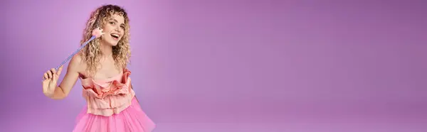 Gaie femme bouclée en robe rose avec baguette magique souriant à la caméra, concept de fée des dents, bannière — Photo de stock