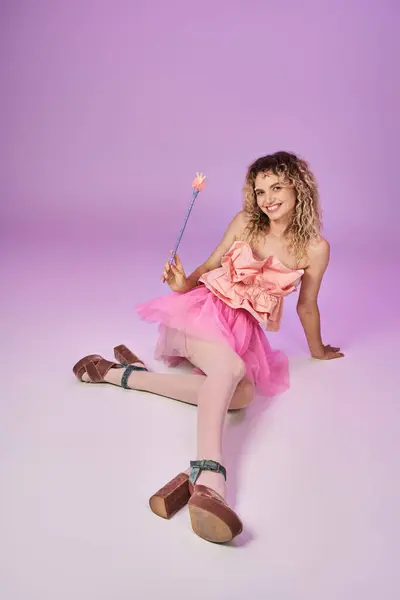 Fada de dente encaracolado bonita em vestido rosa com varinha mágica sentado no chão e posando em pano de fundo rosa — Fotografia de Stock