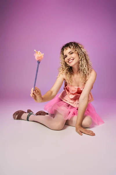 Joyful curly woman in festive pink dress sitting on floor holding magic wand, tooth fairy concept — Stock Photo