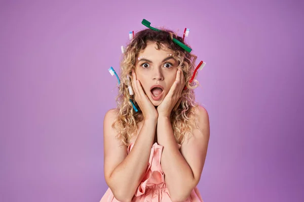 Schockierte Frau in rosa Kleid und Zahnbürsten im Haar, die Hände zu Wangen erhoben, Zahnfee-Konzept — Stockfoto