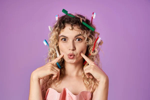Joyous tooth fairy with toothbrushes in her hair pointing fingers at her cheeks looking at camera — Stock Photo