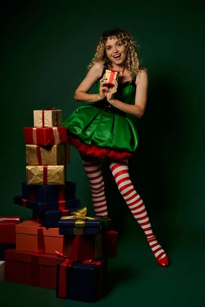 Mulher alegre em traje de elfo verde em pé perto de pilha de presentes e mostrando um presente na câmera — Fotografia de Stock