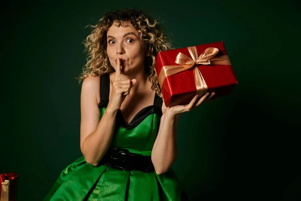 Pretty elf posing in green dress with present in hands on green backdrop showing silence gesture — Stock Photo