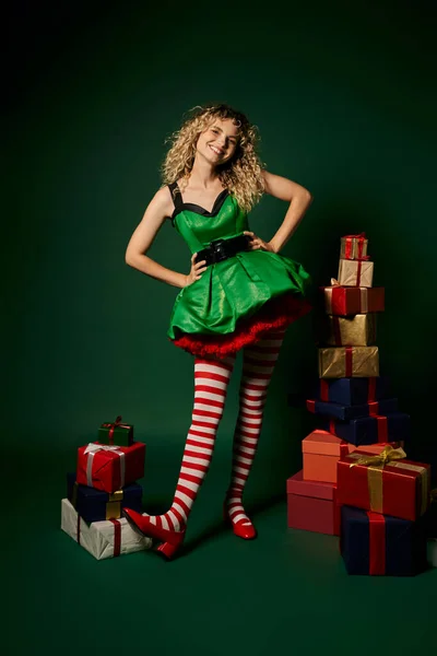 Joyful woman in elf costume posing with arms akimbo next to pile of presents on green background — Stock Photo