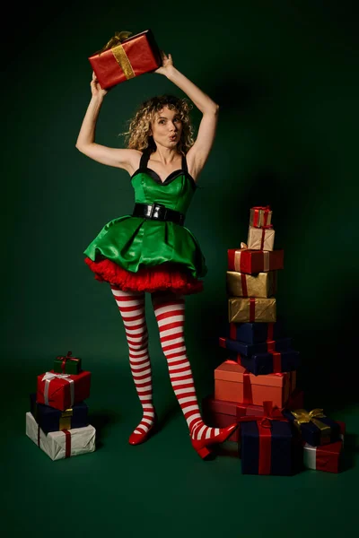 Femme étonnée en robe verte tenant cadeau au-dessus de la pile de cadeaux, concept elfe nouvelle année — Photo de stock