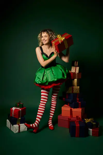 Nouvel an bouclé elfe debout près pile de cadeaux et mystérieusement montrant l'un d'eux à la caméra — Photo de stock