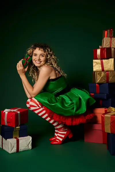 Веселая женщина в зеленом платье на корточках возле подарков на зеленом фоне, новогодняя концепция эльфа — стоковое фото