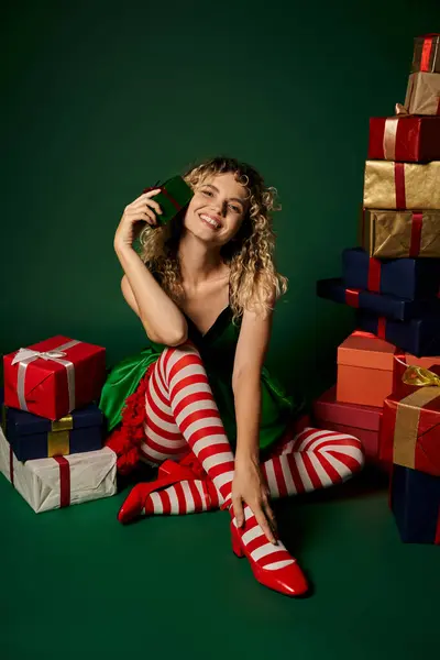 Gai nouvel an elfe en robe verte assis sur le sol avec les jambes croisées entourées de cadeaux — Photo de stock