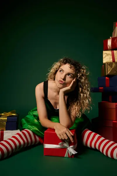 Sonhador ano novo elfo com lábios despojados em vestido verde sentado no chão com presentes olhando para longe — Fotografia de Stock