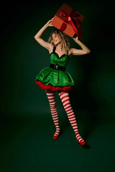 Smiley nouvel an elfe en robe verte festive tenant présent au-dessus de sa tête et détournant les yeux — Photo de stock
