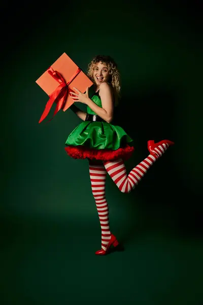 Щасливий кучерявий новорічний ельф стоїть на одній нозі і тримає величезний подарунок в руках посміхається на камеру — стокове фото