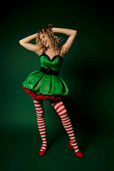 Heureux nouvel elfe de l'année en robe verte posant avec les bras levés cheveux touchants sur fond vert foncé — Photo de stock