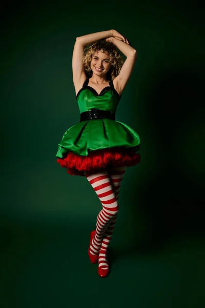 Femme gaie habillée comme elfe de nouvelle année posant avec son bras levé aux cheveux et souriant à la caméra — Photo de stock