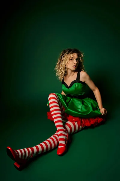 Dreamy curly woman dressed as new year elf sitting on floor posing on dark green background — Stock Photo