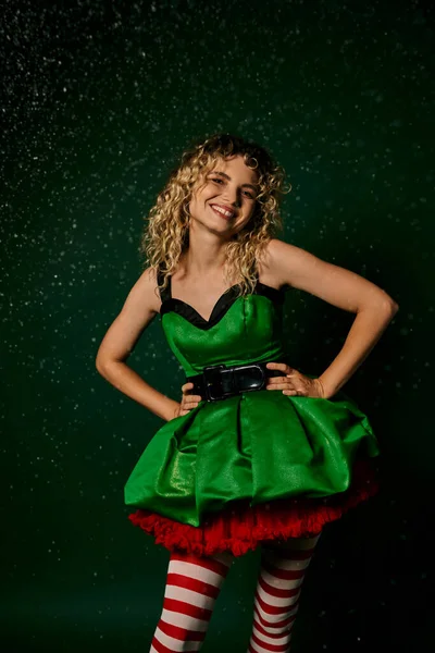 Femme gaie en robe de fête verte souriant et tenant la main akimbo, concept elfe nouvelle année — Photo de stock