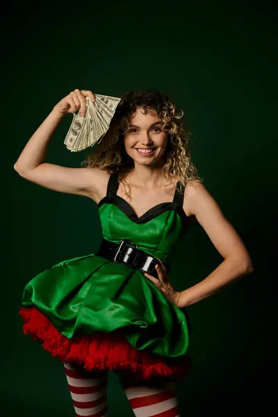 Heureuse femme bouclée habillée comme elfe de nouvelle année avec un bras akimbo et avec de l'argent dans une autre main — Photo de stock