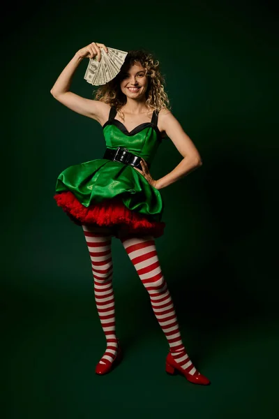 Cheerful new year elf in green festive dress and stockings holding money with one arm akimbo — Stock Photo