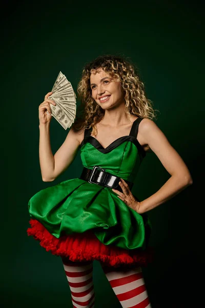 Счастливая блондинка в зеленом платье улыбается и держит деньги с одной рукой на бедре, новый год эльф концепции — стоковое фото